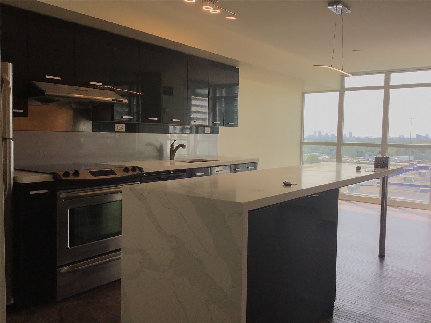 This North York condo renovation features a modern kitchen with a large quartz island, sleek dark cabinetry, and stainless steel appliances. Floor-to-ceiling windows bring in natural light and city views, creating a clean, contemporary space designed for condo living and everyday functionality.