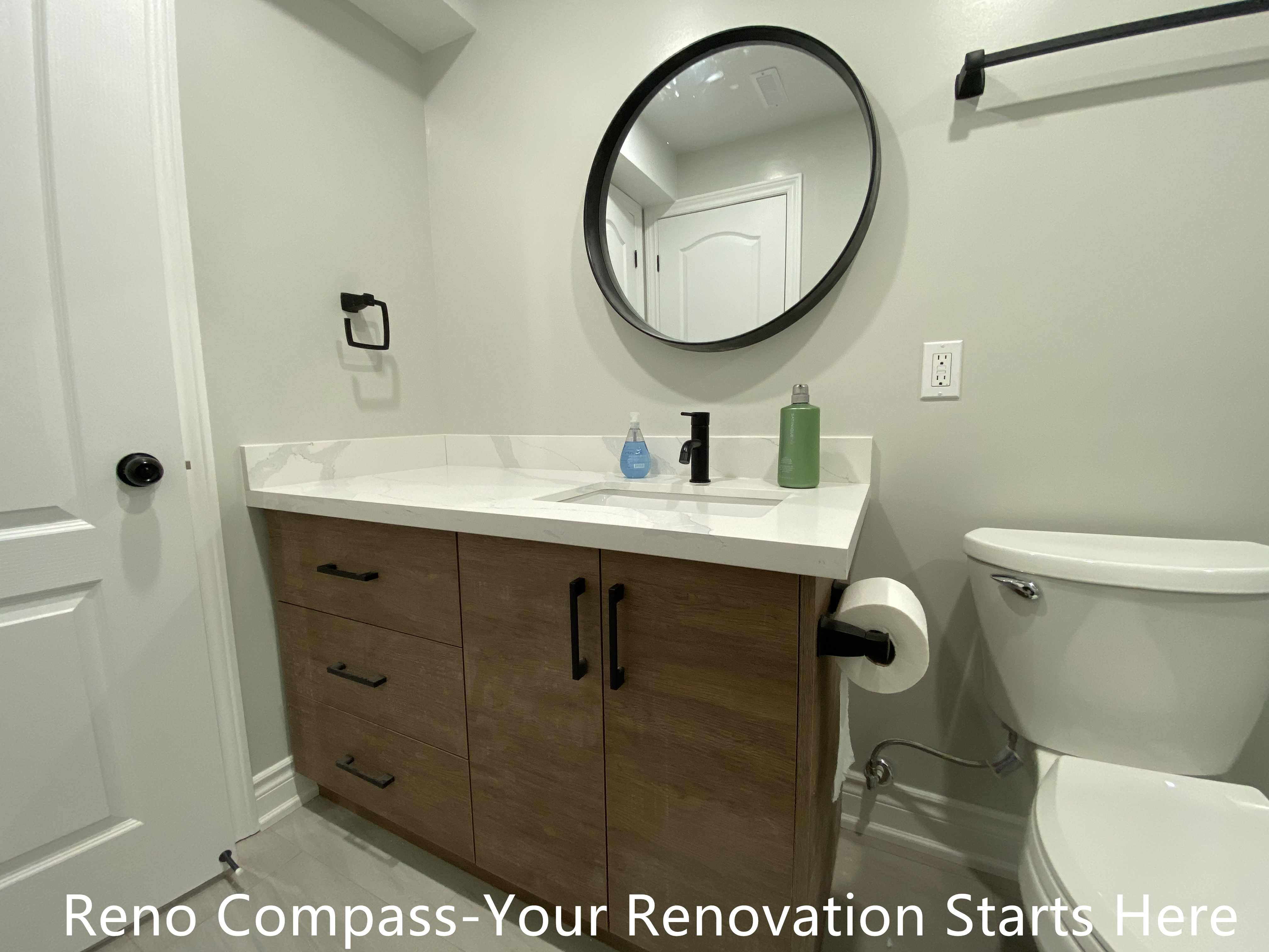 This Aurora basement bathroom renovation showcases a custom wood-finish vanity paired with a white quartz countertop, combining durability with a warm, modern aesthetic. Matte black hardware, a round framed mirror, and minimalist fixtures create a clean and balanced look while maintaining everyday practicality. The layout is optimized for basement conditions, offering ample storage and easy maintenance without compromising style. This bathroom upgrade enhances both functionality and long-term value as part of a fully finished basement renovation completed by Reno Compass.