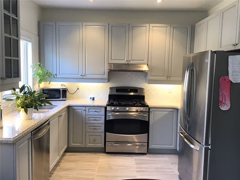 This Woodbridge interior renovation features a clean and modern kitchen design with grey shaker cabinets, quartz countertops, and stainless steel appliances. The layout maximizes functionality with an L-shaped counter, built-in dishwasher, and bright natural lighting, creating a practical yet stylish cooking space for everyday living.
