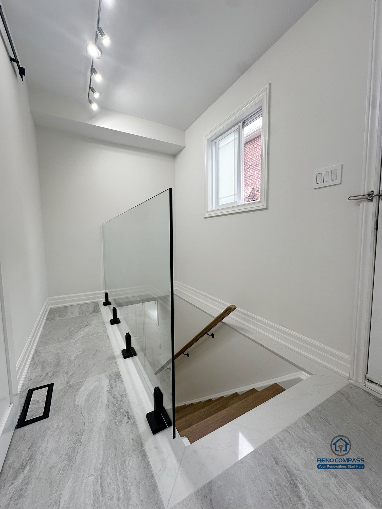 This image highlights a completed basement staircase renovation in Pickering, featuring a clean and modern stair design with a glass guardrail for visual openness and safety. The stair landing is finished with durable tile flooring, while the glass railing allows light to pass through the space, creating a brighter and more connected transition between levels. Track lighting and neutral wall finishes enhance clarity and usability, making the stair area both functional and visually consistent with the rest of the basement renovation.