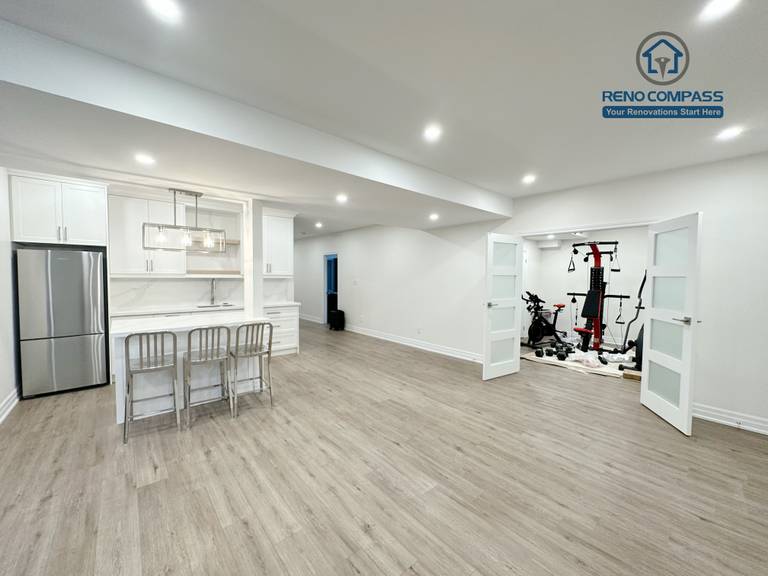 This image showcases an open-concept basement renovation in Bradford, featuring a custom bar kitchen with white cabinetry, quartz countertops, and integrated lighting. The central island provides both dining and entertaining functionality, seamlessly connecting the kitchen to the living area. Designed and completed by a trusted Toronto renovation company, this basement renovation enhances usability, brightness, and long-term comfort for the home.