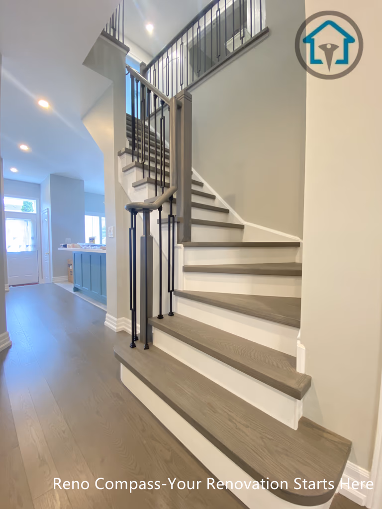 This modern staircase renovation features warm wood treads paired with crisp white risers and slim black metal railings, creating a clean and open visual transition between floors. The design improves circulation, sightlines, and natural light flow while maintaining safety and structural clarity. Carefully coordinated finishes allow the staircase to integrate seamlessly with the surrounding flooring and kitchen area, delivering a cohesive interior renovation by Reno Compass.