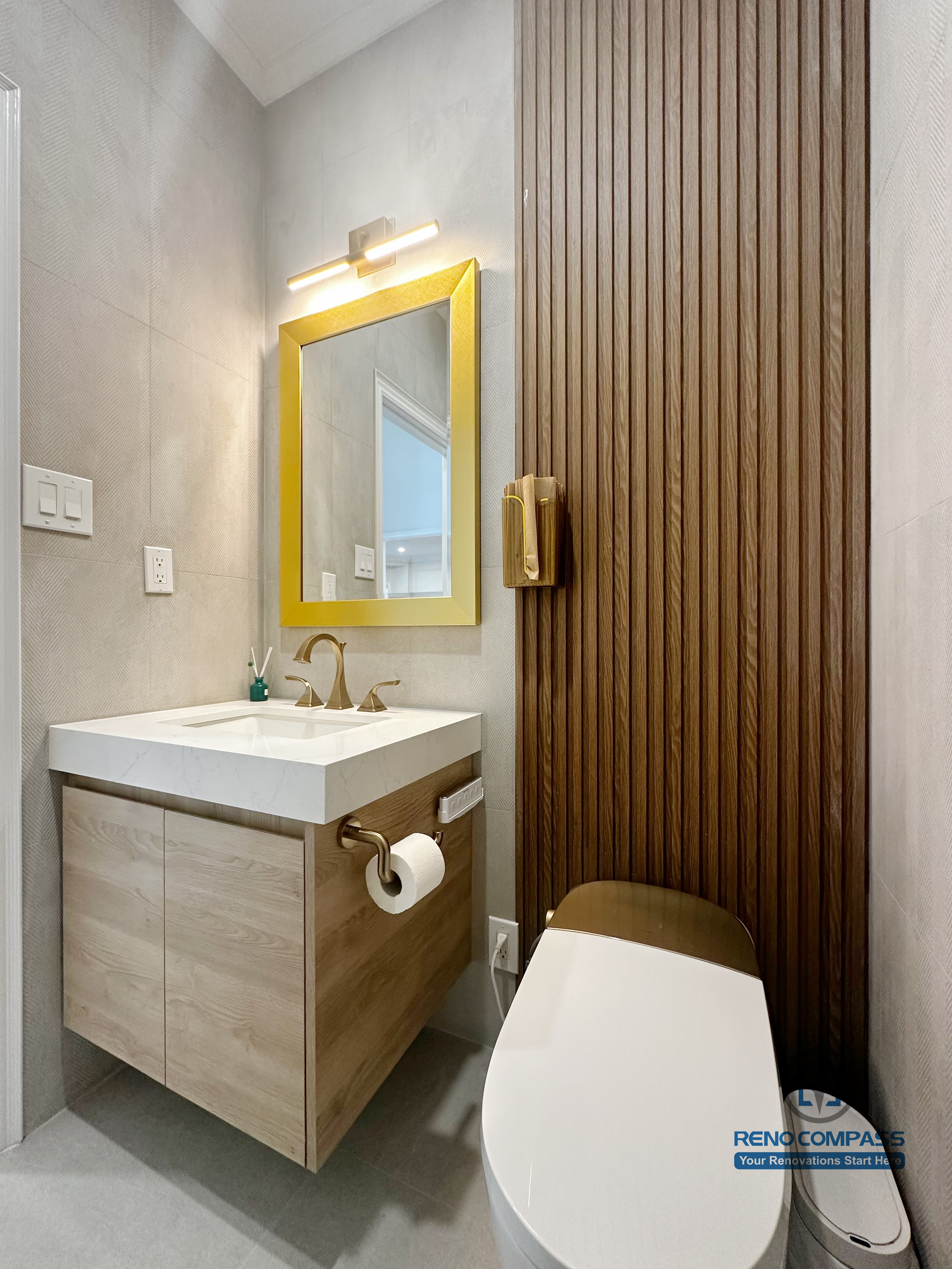 This image showcases a refined bathroom renovation detail completed in North York, featuring a floating wood vanity with integrated storage, a modern rectangular sink, and brushed gold fixtures. A gold-framed mirror paired with warm lighting enhances the visual balance, while the vertical wood accent wall adds texture and depth to the space. This design highlights careful material coordination and precise installation, creating a functional and elegant bathroom layout suited for contemporary North York homes.