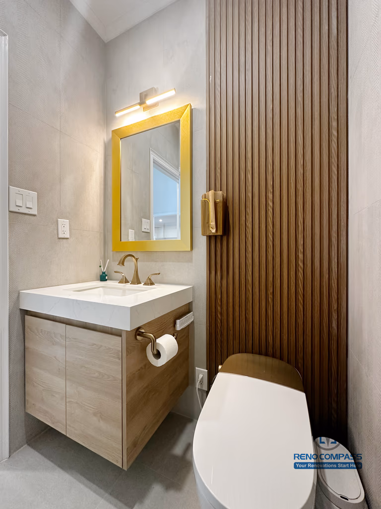 This image showcases a refined bathroom renovation detail completed in North York, featuring a floating wood vanity with integrated storage, a modern rectangular sink, and brushed gold fixtures. A gold-framed mirror paired with warm lighting enhances the visual balance, while the vertical wood accent wall adds texture and depth to the space. This design highlights careful material coordination and precise installation, creating a functional and elegant bathroom layout suited for contemporary North York homes.