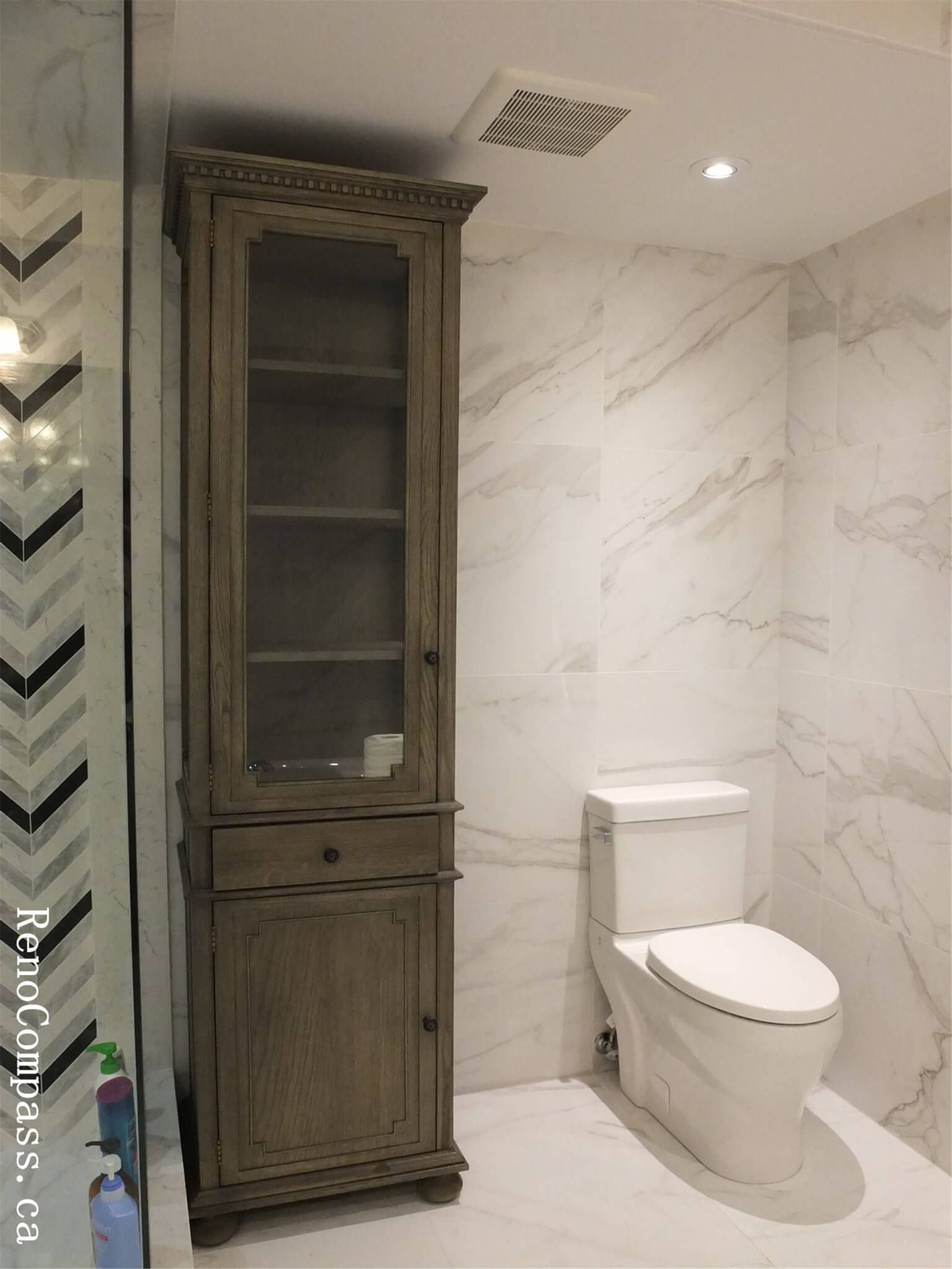 A tall, rustic grey wood linen tower cabinet next to a modern one-piece toilet and marble wall tiles.