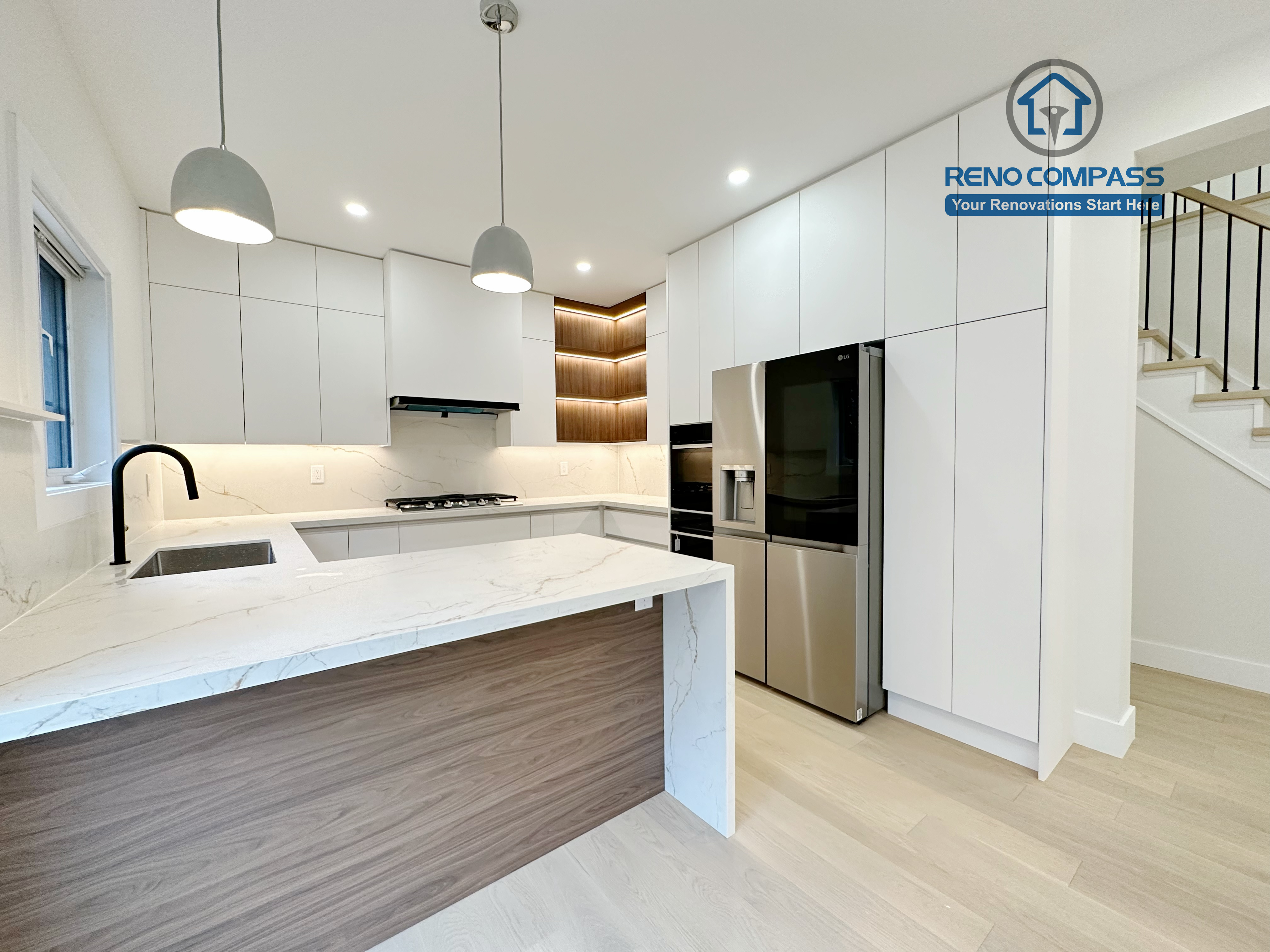 This image presents a modern kitchen renovation in Richmond Hill featuring full-height custom white cabinetry, a quartz waterfall island with wood panel detailing, and integrated stainless steel appliances. The design includes a built-in gas cooktop, minimalist pendant lights, recessed ceiling lighting, and light hardwood flooring for a clean, contemporary look. Large windows and patio access enhance natural light and openness. Completed by a professional Toronto Renovation Company, this kitchen renovation reflects refined craftsmanship, efficient layout planning, and modern family-focused design.