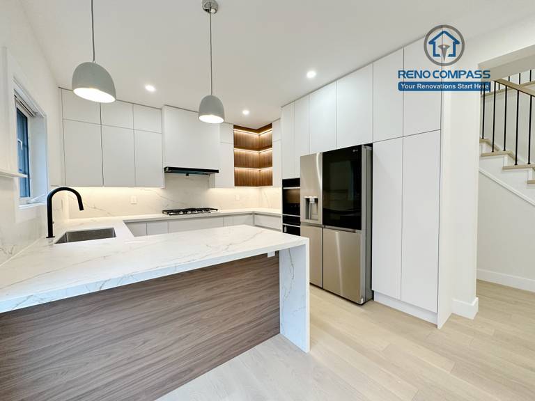 This image presents a modern kitchen renovation in Richmond Hill featuring full-height custom white cabinetry, a quartz waterfall island with wood panel detailing, and integrated stainless steel appliances. The design includes a built-in gas cooktop, minimalist pendant lights, recessed ceiling lighting, and light hardwood flooring for a clean, contemporary look. Large windows and patio access enhance natural light and openness. Completed by a professional Toronto Renovation Company, this kitchen renovation reflects refined craftsmanship, efficient layout planning, and modern family-focused design.