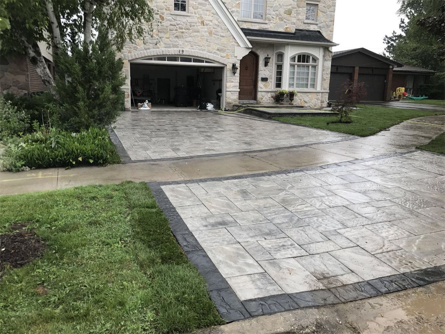 Residential landscaping renovation in Toronto featuring a large interlocking driveway and connecting walkway with decorative borders. The design enhances curb appeal while providing a durable and visually balanced front yard entrance.