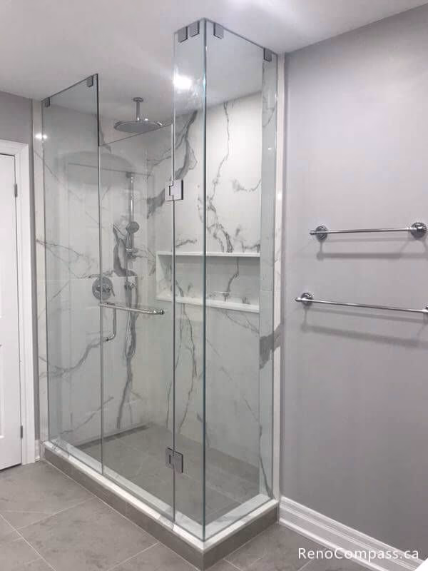This Markham bathroom renovation showcases a spacious frameless glass shower with marble-pattern porcelain tiles and built-in shelving. The space also features a custom white vanity with quartz countertop, modern lighting, and large floor tiles that create a clean and elegant look. The updated layout improves both functionality and visual appeal, delivering a bright and contemporary bathroom design for the homeowner.