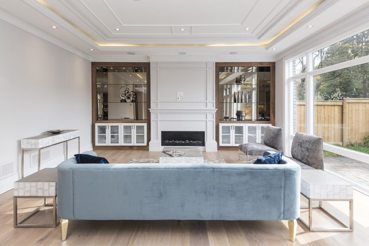 This Newmarket interior renovation showcases a spacious open-concept living room designed by a professional Toronto renovation company. The space features wide plank hardwood flooring, a custom built-in fireplace with elegant wall panel detailing, and symmetrical display cabinetry that enhances functionality and style. Large floor-to-ceiling windows bring in abundant natural light, creating a bright and welcoming environment. The coffered ceiling with recessed LED lighting adds a modern luxury touch, transforming the living area into a refined and comfortable gathering space for contemporary family living in Newmarket and the GTA.