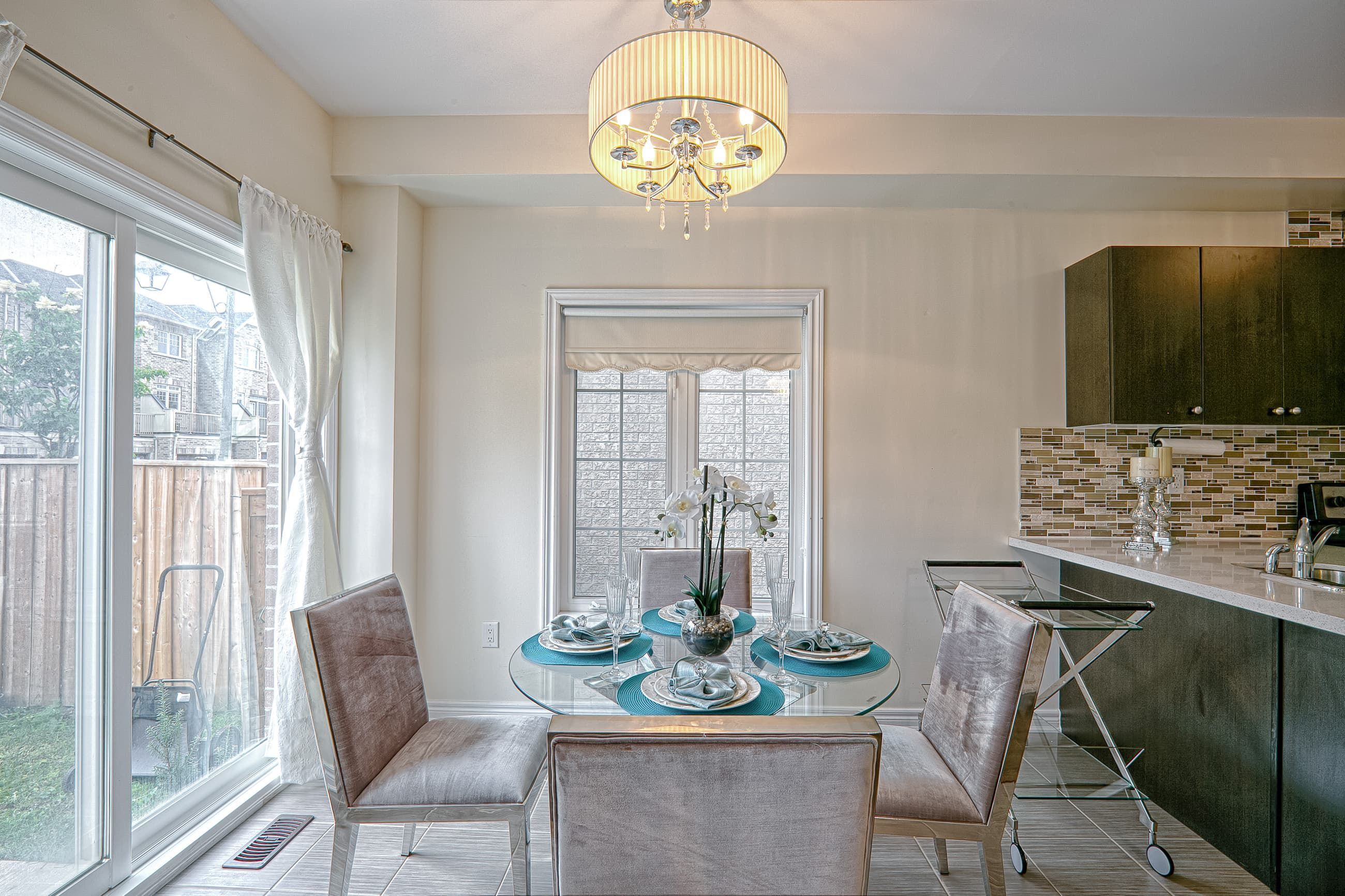This elegant dining space features a modern glass-top dining table, upholstered chairs, and a stylish chandelier that enhances the warm and inviting atmosphere. Large sliding doors allow abundant natural light to fill the space, while the open connection to the contemporary kitchen with mosaic backsplash and dark cabinetry creates a cohesive and functional layout. Designed with both comfort and visual balance in mind, this interior reflects the quality craftsmanship of a professional Toronto renovation company specializing in modern residential upgrades and open-concept living design.