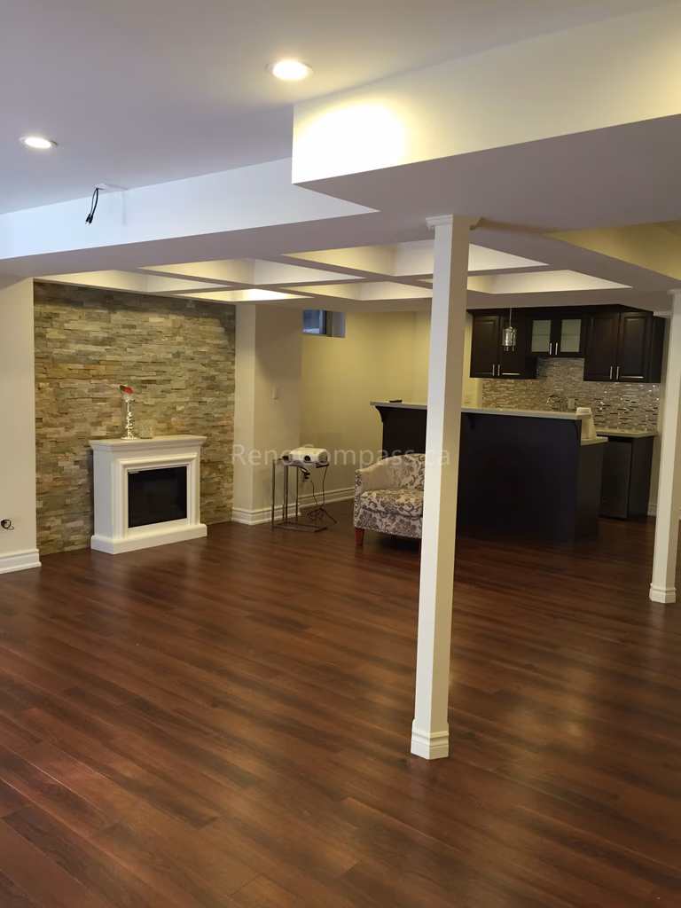Open-concept basement renovation in Toronto featuring new hardwood flooring, finished support columns, and a modern stone fireplace feature wall. The upgraded layout includes a functional entertainment area and stylish bar space, creating a warm and inviting environment for family gatherings. Completed by Reno Compass, a trusted Toronto renovation company, this project highlights quality craftsmanship and practical basement living design for modern homes.