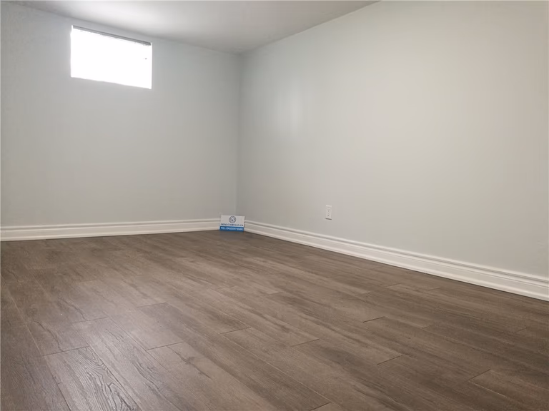 Finished interior space from a Pickering basement renovation project featuring new wood-look flooring, clean white walls, baseboard trim, and a bright window for natural light. This simple and functional room upgrade improves comfort and usability, completed by a professional Toronto renovation company.