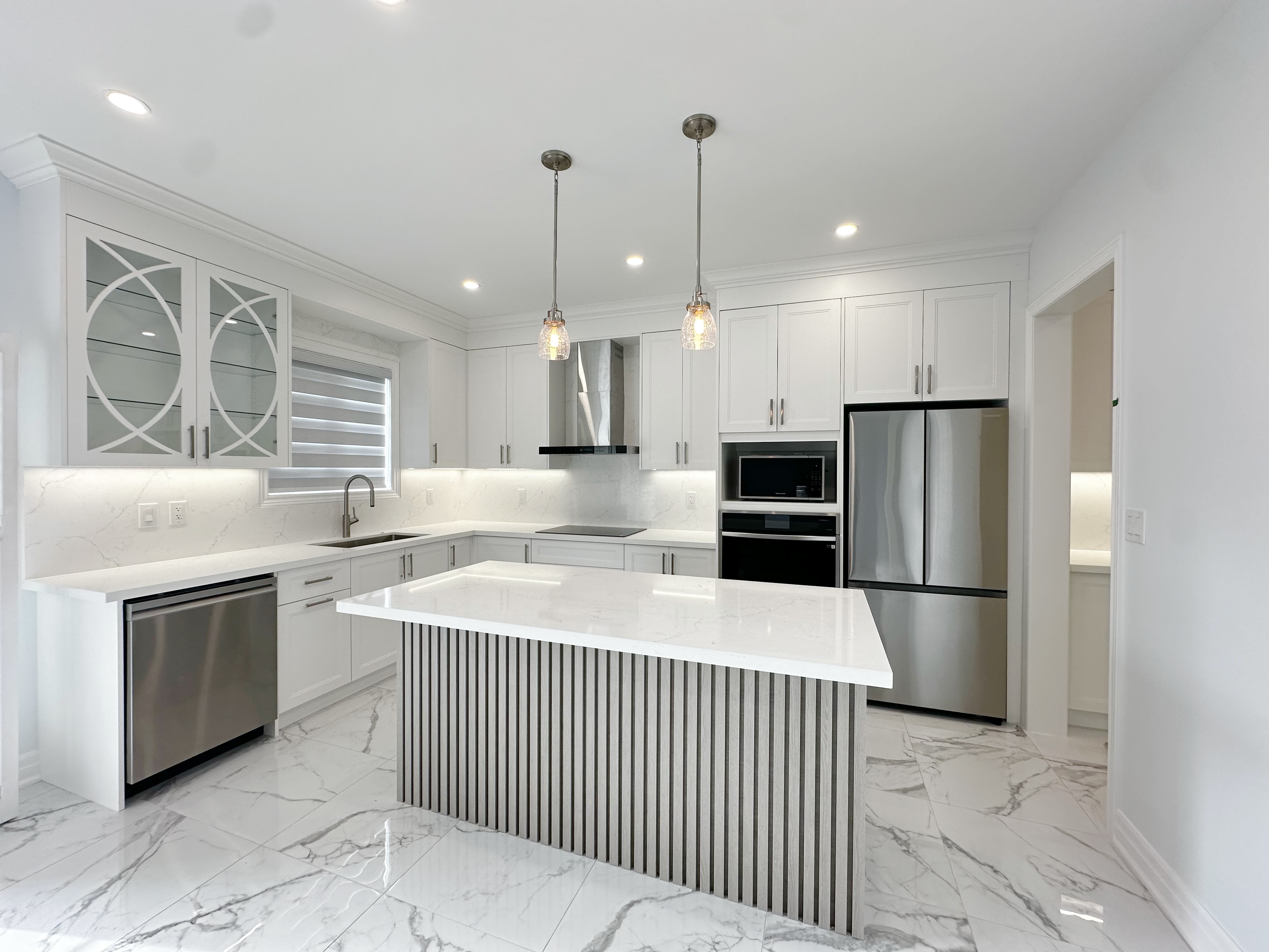 This image showcases a modern kitchen renovation in a Markham house, featuring custom white cabinetry, a large central island with vertical panel detailing, marble-style flooring, and integrated stainless steel appliances. The open layout enhances natural light, functionality, and everyday family use.