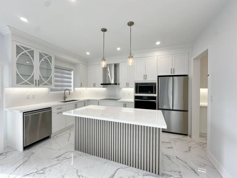 This image showcases a modern kitchen renovation in a Markham house, featuring custom white cabinetry, a large central island with vertical panel detailing, marble-style flooring, and integrated stainless steel appliances. The open layout enhances natural light, functionality, and everyday family use.