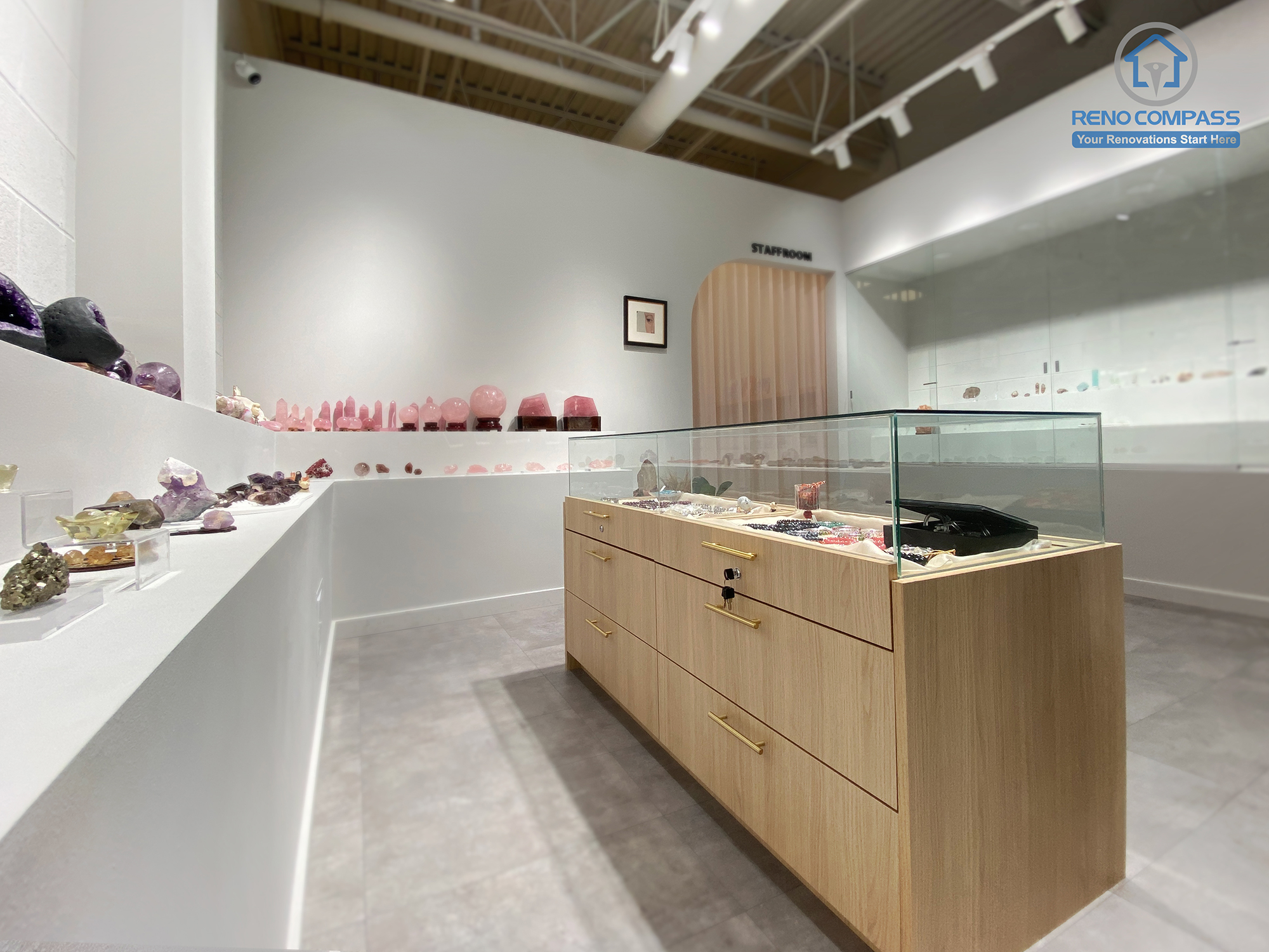 This image captures the main retail area of a crystal shop renovation in Markham, featuring a custom-built wood display island with glass showcase, integrated storage, and refined brass hardware. Surrounding wall-mounted shelves are designed to present crystals in a clean, gallery-style layout. The combination of minimalist white walls, polished concrete flooring, and professional track lighting creates a calm and premium retail environment. As a Toronto renovation company, Reno Compass delivers tailored retail renovations that enhance product presentation and customer flow.