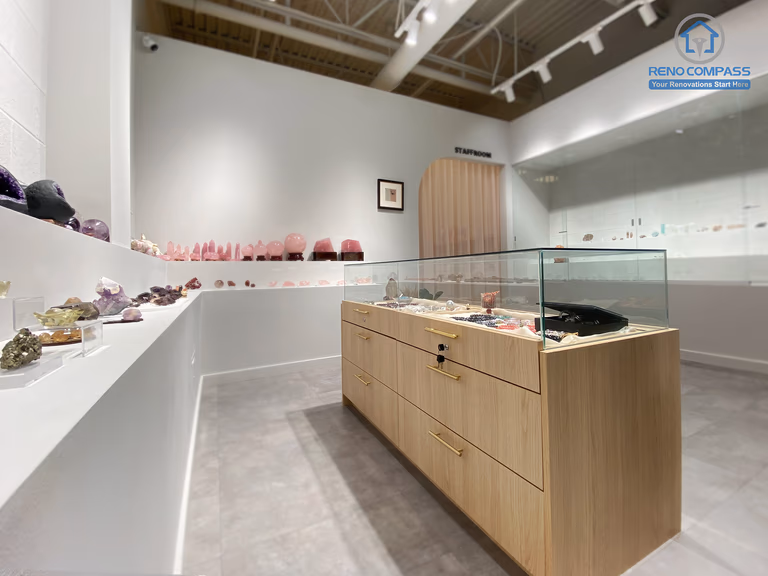 This image captures the main retail area of a crystal shop renovation in Markham, featuring a custom-built wood display island with glass showcase, integrated storage, and refined brass hardware. Surrounding wall-mounted shelves are designed to present crystals in a clean, gallery-style layout. The combination of minimalist white walls, polished concrete flooring, and professional track lighting creates a calm and premium retail environment. As a Toronto renovation company, Reno Compass delivers tailored retail renovations that enhance product presentation and customer flow.