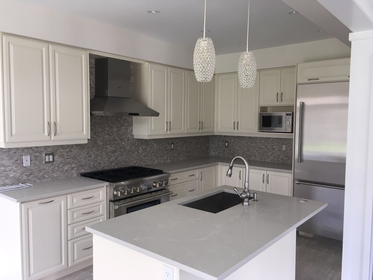 This Toronto interior renovation features a bright and modern kitchen design with elegant white shaker cabinets, a large quartz island with built-in sink, and stylish pendant lighting. The space is enhanced by a mosaic backsplash, stainless steel appliances, and a functional open layout, creating a clean, timeless, and highly practical cooking environment.