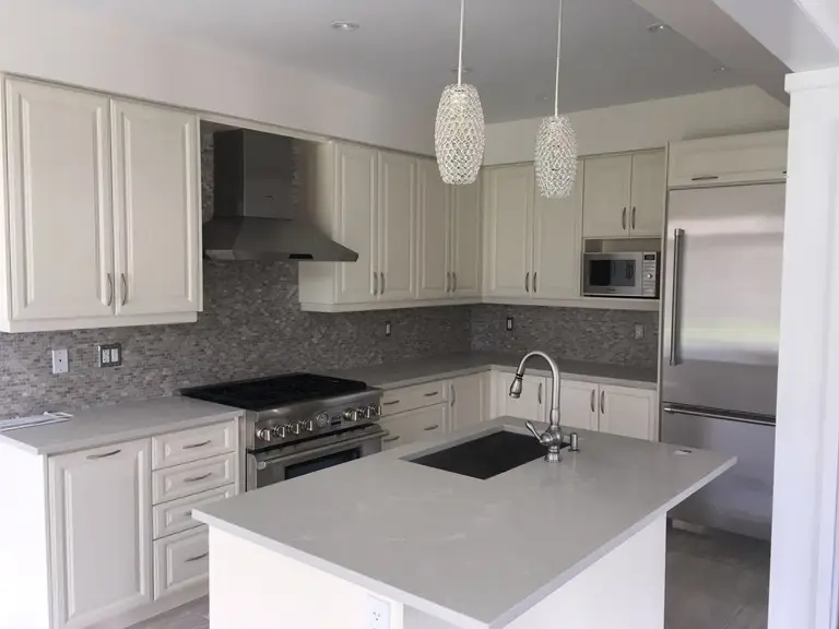 This Toronto interior renovation features a bright and modern kitchen design with elegant white shaker cabinets, a large quartz island with built-in sink, and stylish pendant lighting. The space is enhanced by a mosaic backsplash, stainless steel appliances, and a functional open layout, creating a clean, timeless, and highly practical cooking environment.
