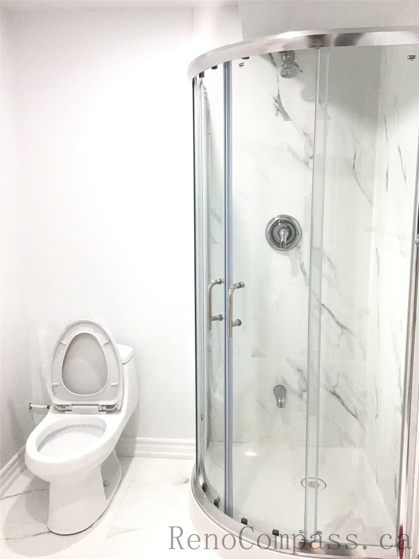 Modern basement bathroom renovation in Pickering featuring a curved glass shower enclosure with marble-style wall panels and a clean white toilet installation. The bright neutral finishes and compact layout maximize functionality while maintaining a sleek contemporary look. Professionally designed and completed by Reno Compass Inc., a trusted Toronto Renovation Company.