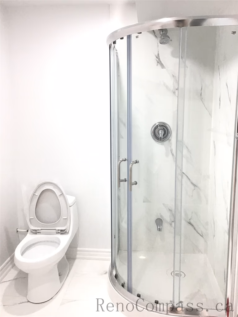 Modern basement bathroom renovation in Pickering featuring a curved glass shower enclosure with marble-style wall panels and a clean white toilet installation. The bright neutral finishes and compact layout maximize functionality while maintaining a sleek contemporary look. Professionally designed and completed by Reno Compass Inc., a trusted Toronto Renovation Company.