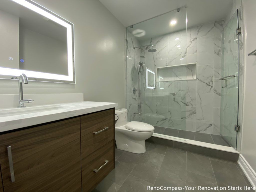 This Oakville bathroom renovation showcases a modern walk-in glass shower with marble-look wall tiles, built-in shower niche, and sleek frameless enclosure for a clean contemporary design. The space also features a custom wood vanity with quartz countertop, LED backlit mirror, and upgraded fixtures that enhance both functionality and visual appeal. Completed by a professional Toronto renovation company, this bathroom upgrade focuses on maximizing brightness, storage, and comfort while delivering a stylish and durable modern bathroom space.