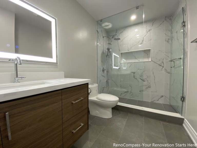 This Oakville bathroom renovation showcases a modern walk-in glass shower with marble-look wall tiles, built-in shower niche, and sleek frameless enclosure for a clean contemporary design. The space also features a custom wood vanity with quartz countertop, LED backlit mirror, and upgraded fixtures that enhance both functionality and visual appeal. Completed by a professional Toronto renovation company, this bathroom upgrade focuses on maximizing brightness, storage, and comfort while delivering a stylish and durable modern bathroom space.