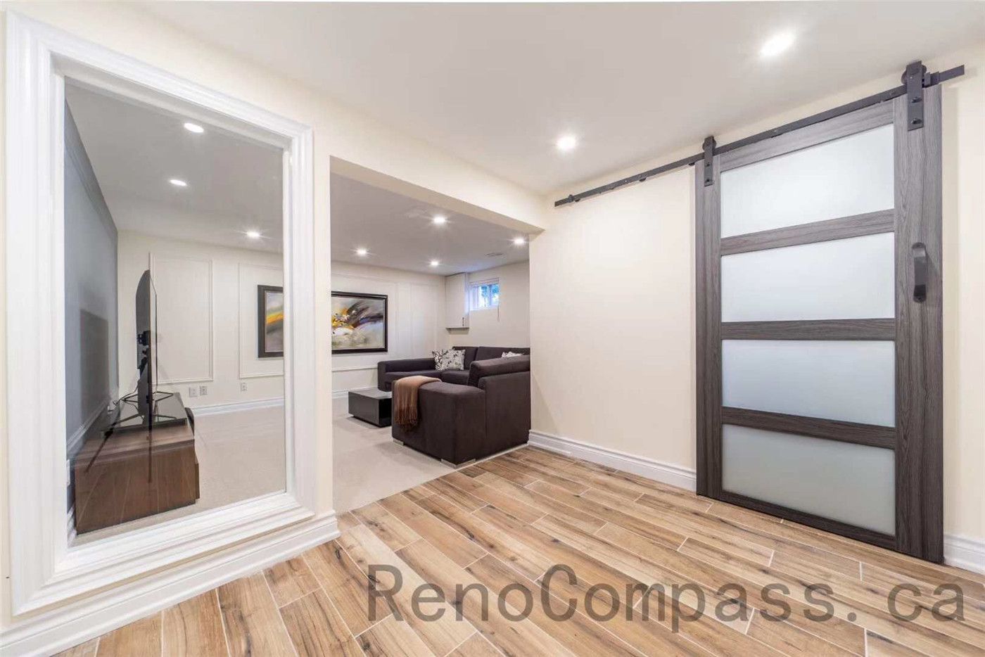This North York basement renovation showcases a modern and functional living space featuring wood-look tile flooring, recessed pot lights, and a stylish sliding barn door with frosted glass panels. The layout connects a cozy entertainment room and lounge area, creating a comfortable environment for family relaxation and movie nights. Completed by a professional Toronto renovation company, the project highlights clean finishes, warm textures, and thoughtful interior design that transforms the basement into a bright and inviting living space.