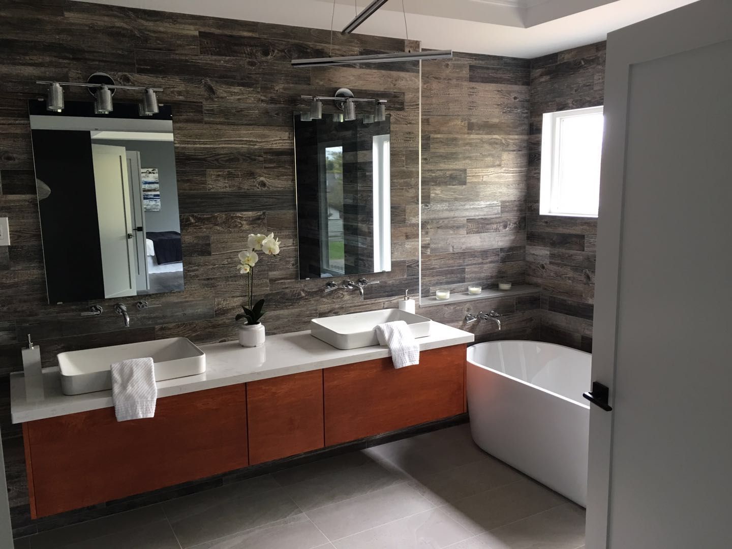 This Toronto interior renovation project showcases a modern bathroom transformation featuring a floating wood vanity with double vessel sinks, sleek wall-mounted mirrors, and a freestanding soaking tub. The textured wood accent wall adds warmth and depth, while large-format flooring enhances the clean, contemporary aesthetic. Designed for both functionality and luxury, this bathroom renovation reflects high-quality craftsmanship and modern interior styling by a professional Toronto renovation company.