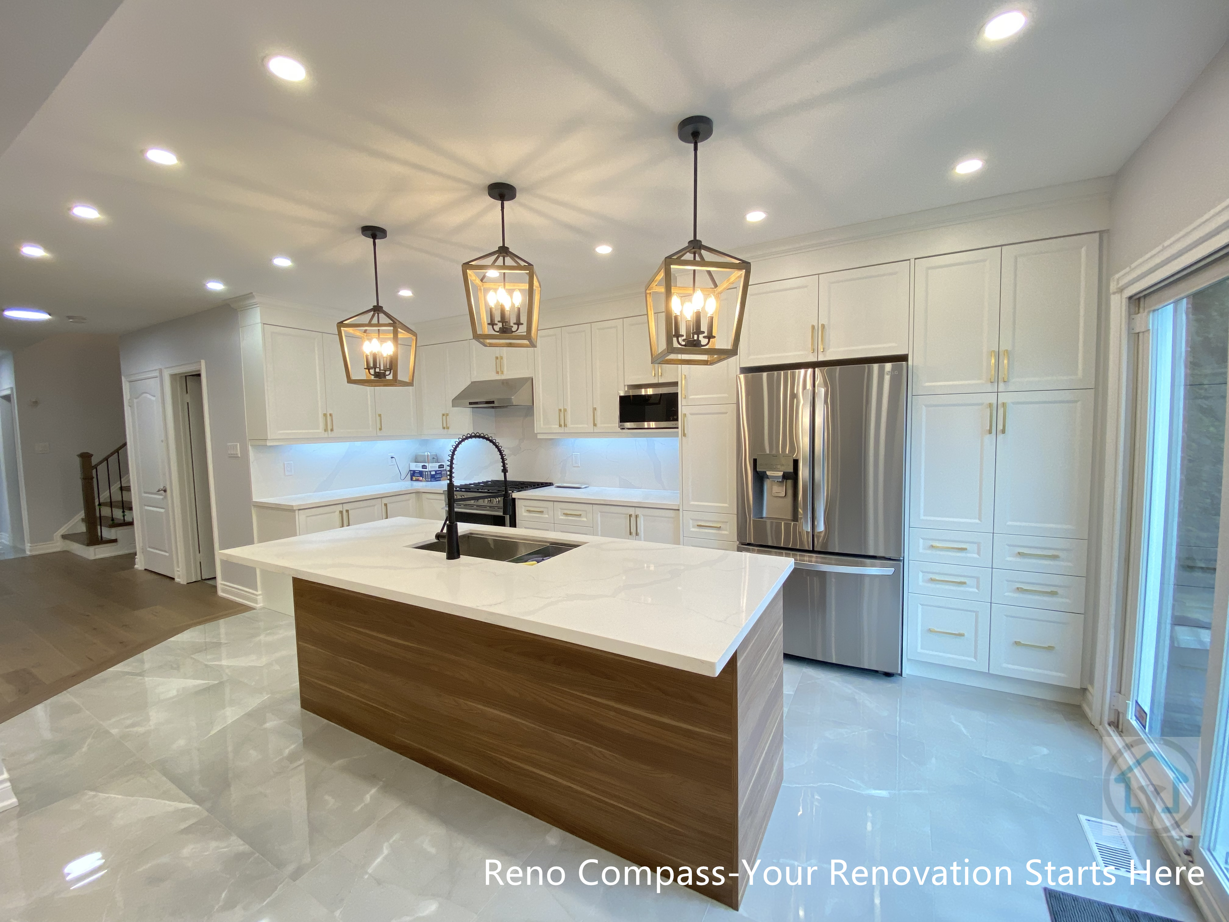 This image highlights a Newmarket interior renovation featuring a long quartz kitchen island with warm wood base cabinetry, designed to anchor the open-concept layout. The linear arrangement of lantern-style pendant lights creates strong visual rhythm and enhances the sense of depth within the space. White shaker cabinets, integrated stainless steel appliances, and glossy marble-look flooring work together to reflect light and elevate the overall brightness. A high-arc faucet and built-in sink add both functionality and modern appeal. Completed by a Toronto renovation company, this kitchen renovation emphasizes clean lines, balanced proportions, and seamless flow between cooking, dining, and outdoor access.