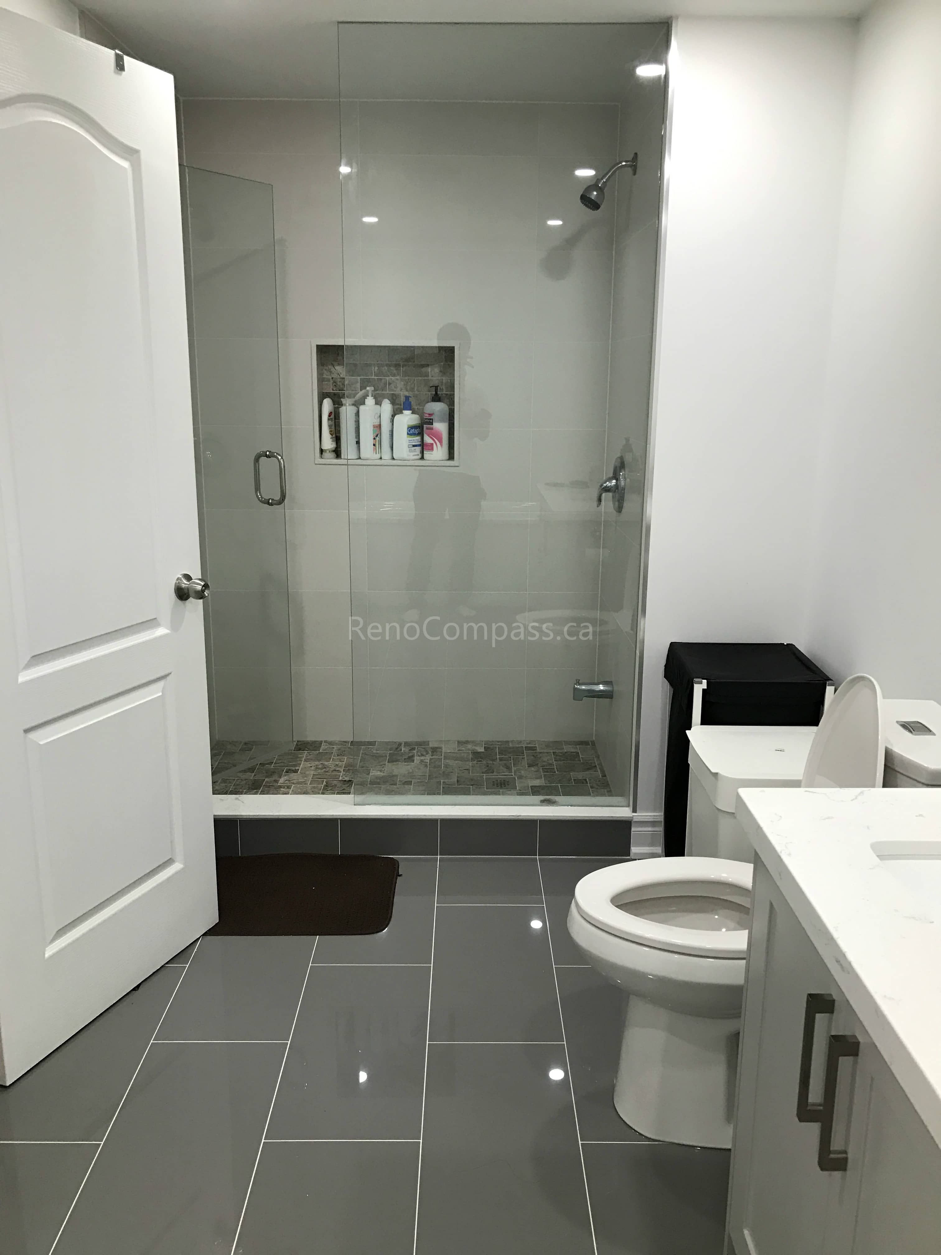 This Toronto bathroom renovation by Reno Compass features a sleek walk-in glass shower with a built-in wall niche for convenient storage and a clean, modern look. Large-format wall tiles and dark floor tiles create a stylish contrast while improving durability and easy maintenance. The upgraded vanity, toilet, and lighting enhance functionality and comfort, delivering a contemporary bathroom space designed for everyday use and long-term value.