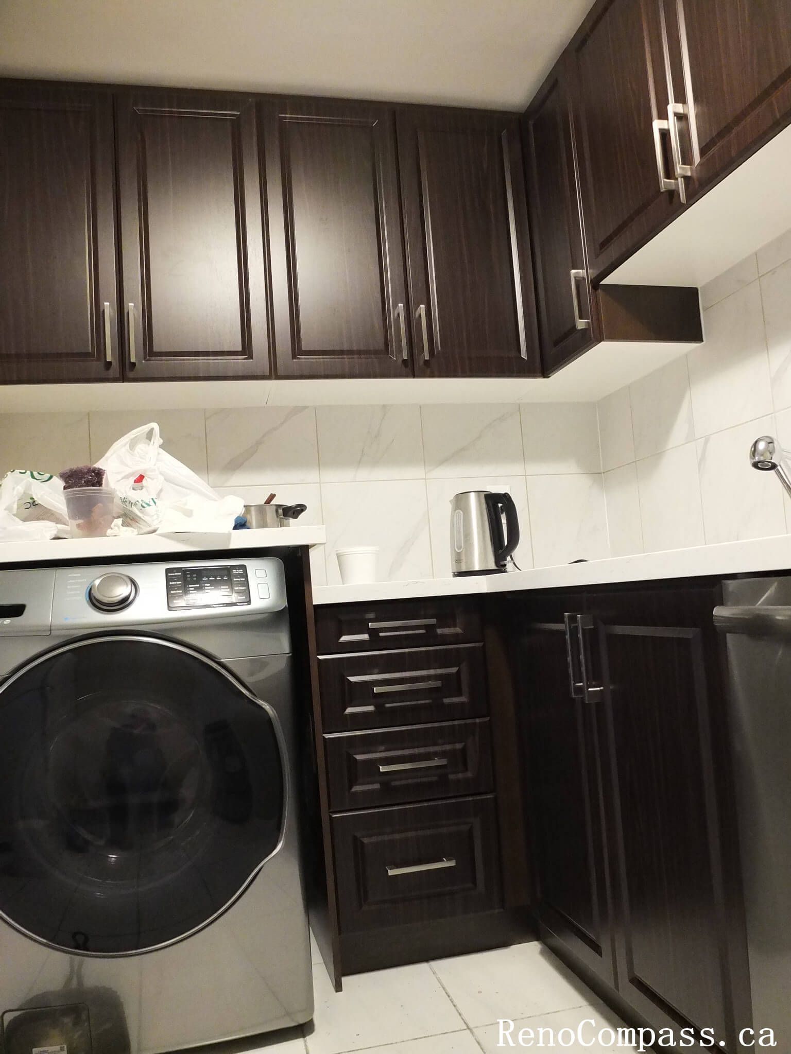 Smart space-saving kitchen design in Mississauga. We integrated a modern front-load washer into the custom dark wood cabinetry to create a highly functional, multi-purpose laundry and kitchen area.