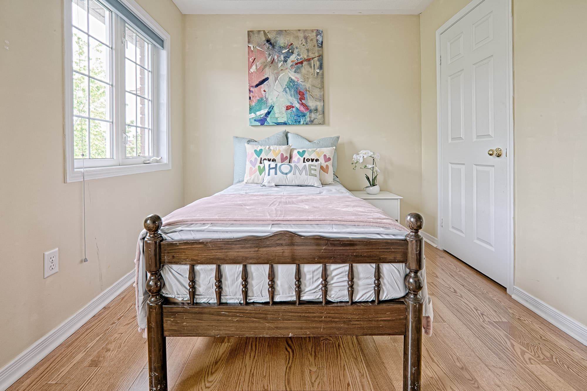 This cozy bedroom showcases a simple and lovely interior design with warm hardwood flooring, soft neutral wall colors, and abundant natural light from large windows. The wooden bed frame paired with light bedding creates a calm and comfortable atmosphere, while minimal decor enhances the sense of space. This design emphasizes practicality, brightness, and everyday comfort, making it ideal for modern family homes and rental properties.