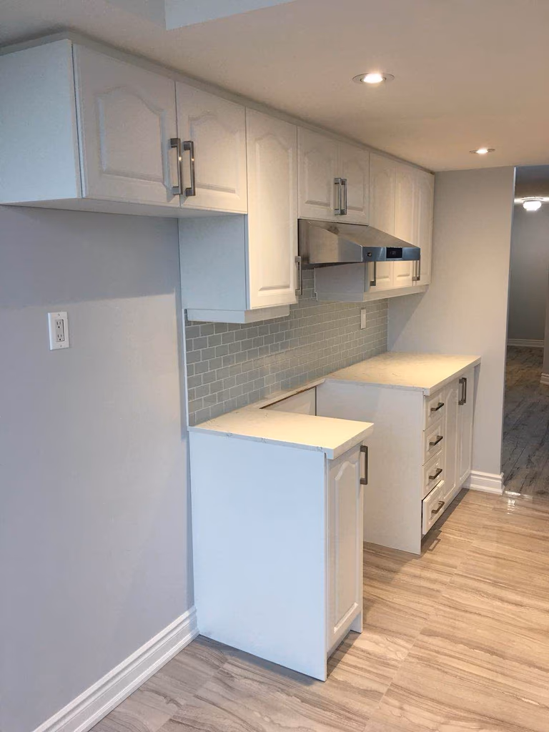 This Toronto interior renovation showcases a clean and modern kitchen design featuring elegant white shaker-style cabinets, a light blue subway tile backsplash, and a durable quartz countertop. The renovation enhances both functionality and visual appeal with integrated sink installation, sleek cabinet hardware, and improved lighting. The bright layout and contemporary finishes create a fresh, welcoming space suitable for modern urban living in Toronto homes.