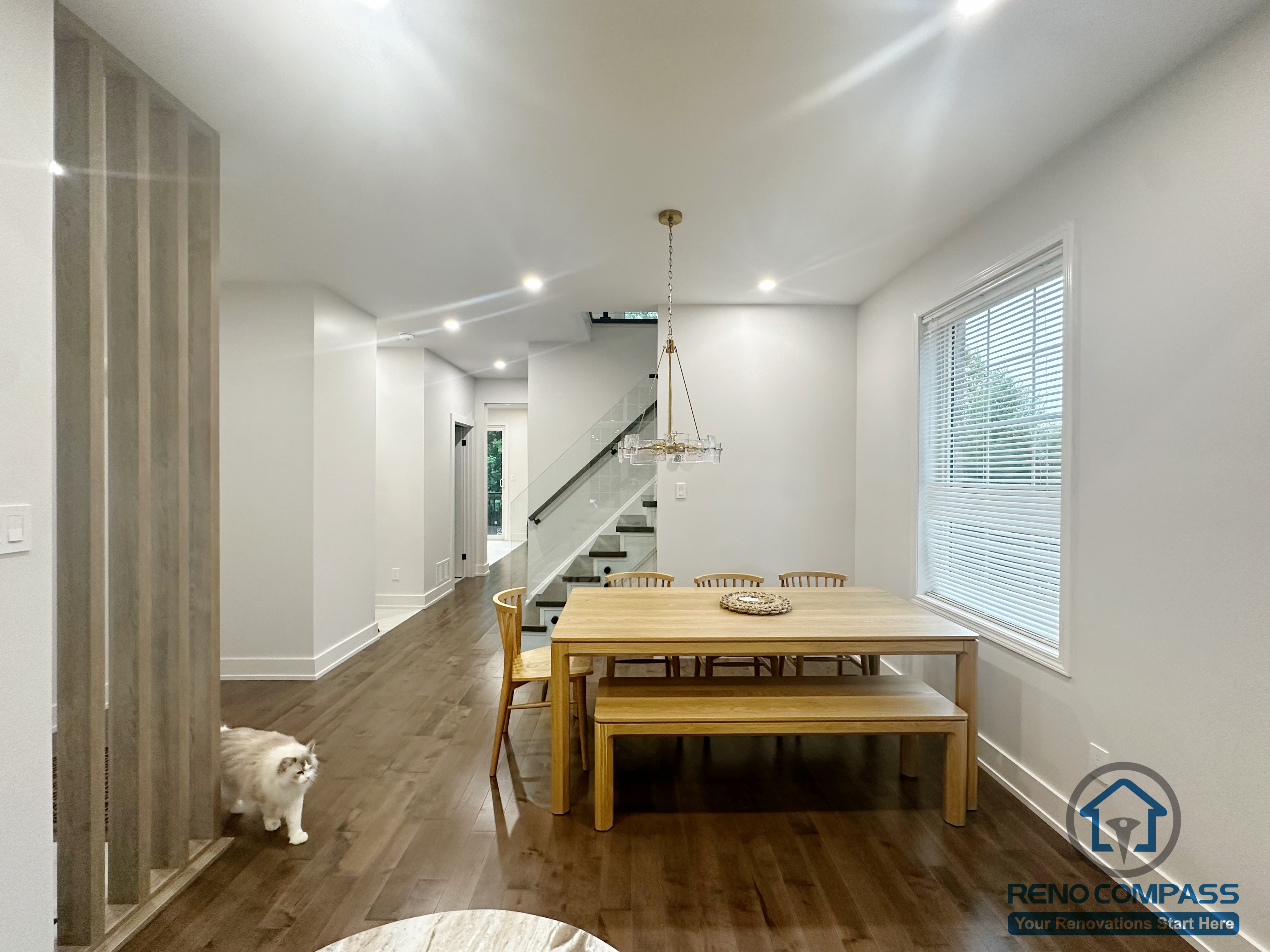 This Oakville interior renovation showcases an open-concept dining area designed for everyday family use and smooth circulation. Natural wood flooring extends throughout the space, creating visual continuity between the dining area, hallway, and staircase. The minimalist dining setup is paired with clean wall finishes and recessed lighting, while the modern staircase with glass railing adds a sense of openness without interrupting sightlines. Large windows introduce natural light, enhancing the warm and balanced atmosphere. The overall layout prioritizes functionality, comfort, and long-term livability within a modern home setting.