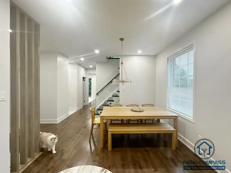 This Oakville interior renovation showcases an open-concept dining area designed for everyday family use and smooth circulation. Natural wood flooring extends throughout the space, creating visual continuity between the dining area, hallway, and staircase. The minimalist dining setup is paired with clean wall finishes and recessed lighting, while the modern staircase with glass railing adds a sense of openness without interrupting sightlines. Large windows introduce natural light, enhancing the warm and balanced atmosphere. The overall layout prioritizes functionality, comfort, and long-term livability within a modern home setting.