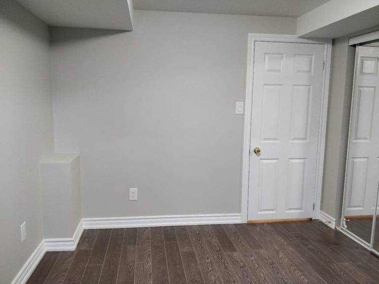 This image features a finished basement room from a Scarborough renovation project, showcasing newly installed dark hardwood flooring, fresh neutral wall paint, and upgraded baseboards for a clean, modern appearance. The space includes a standard entry door and mirrored closet doors, creating a functional layout suitable for a bedroom, home office, or guest suite. The crisp trim detailing and cohesive color palette enhance the overall brightness and comfort of the basement. Completed by Reno Compass Inc., this renovation highlights quality finishes and thoughtful interior upgrades.