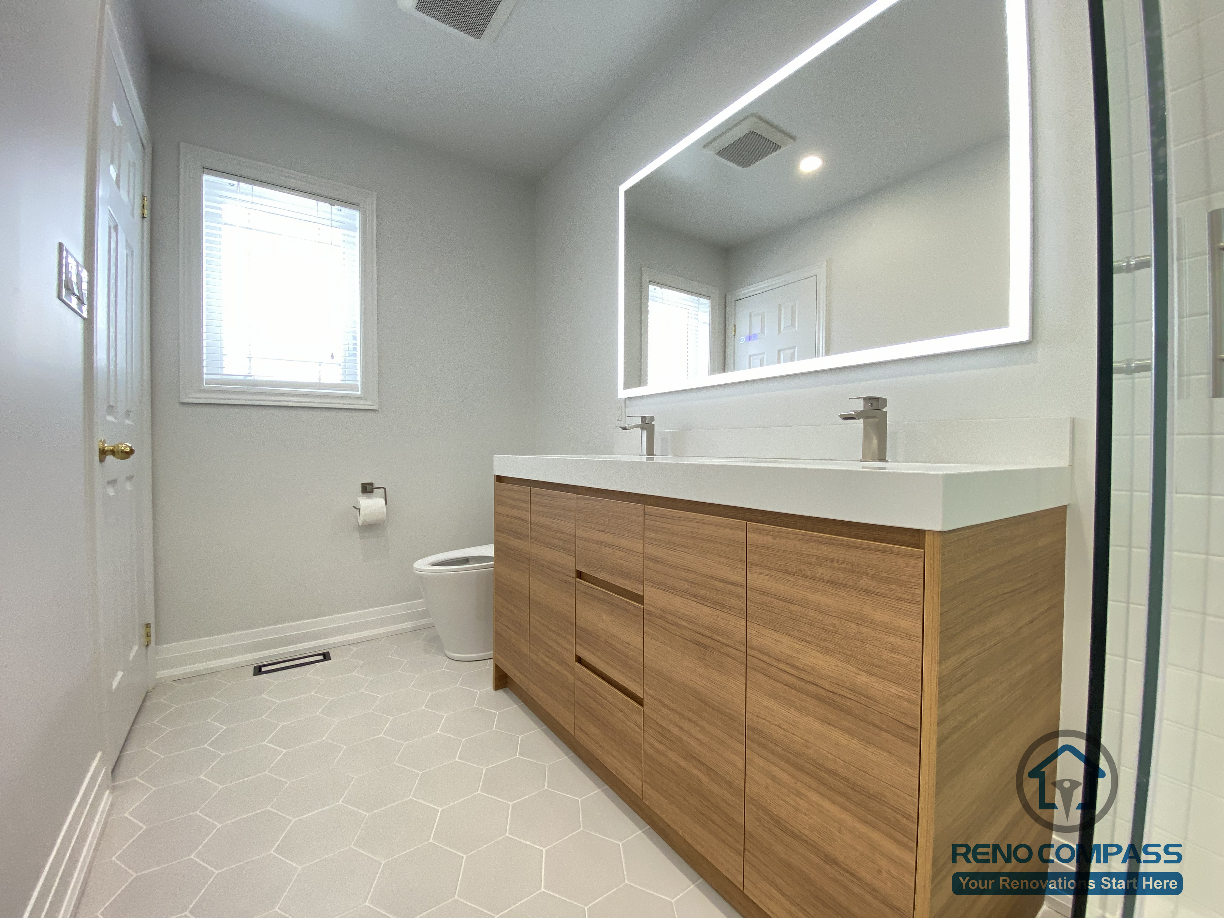This Markham bathroom renovation highlights a custom double sink vanity designed to improve daily efficiency and long-term usability. The wood-tone vanity provides ample concealed storage while adding warmth and visual balance to the space. A full-width LED backlit mirror delivers even, glare-free lighting, enhancing functionality for everyday routines. Slip-resistant hexagon tile flooring improves safety and durability in a high-moisture environment, while neutral wall tones maintain a clean, timeless appearance. Completed by a Toronto renovation company, Reno Compass Inc., this bathroom renovation reflects careful space planning, material coordination, and a modern, practical design approach for Markham homes.