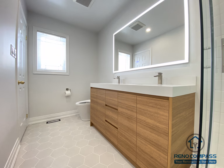 This Markham bathroom renovation highlights a custom double sink vanity designed to improve daily efficiency and long-term usability. The wood-tone vanity provides ample concealed storage while adding warmth and visual balance to the space. A full-width LED backlit mirror delivers even, glare-free lighting, enhancing functionality for everyday routines. Slip-resistant hexagon tile flooring improves safety and durability in a high-moisture environment, while neutral wall tones maintain a clean, timeless appearance. Completed by a Toronto renovation company, Reno Compass Inc., this bathroom renovation reflects careful space planning, material coordination, and a modern, practical design approach for Markham homes.