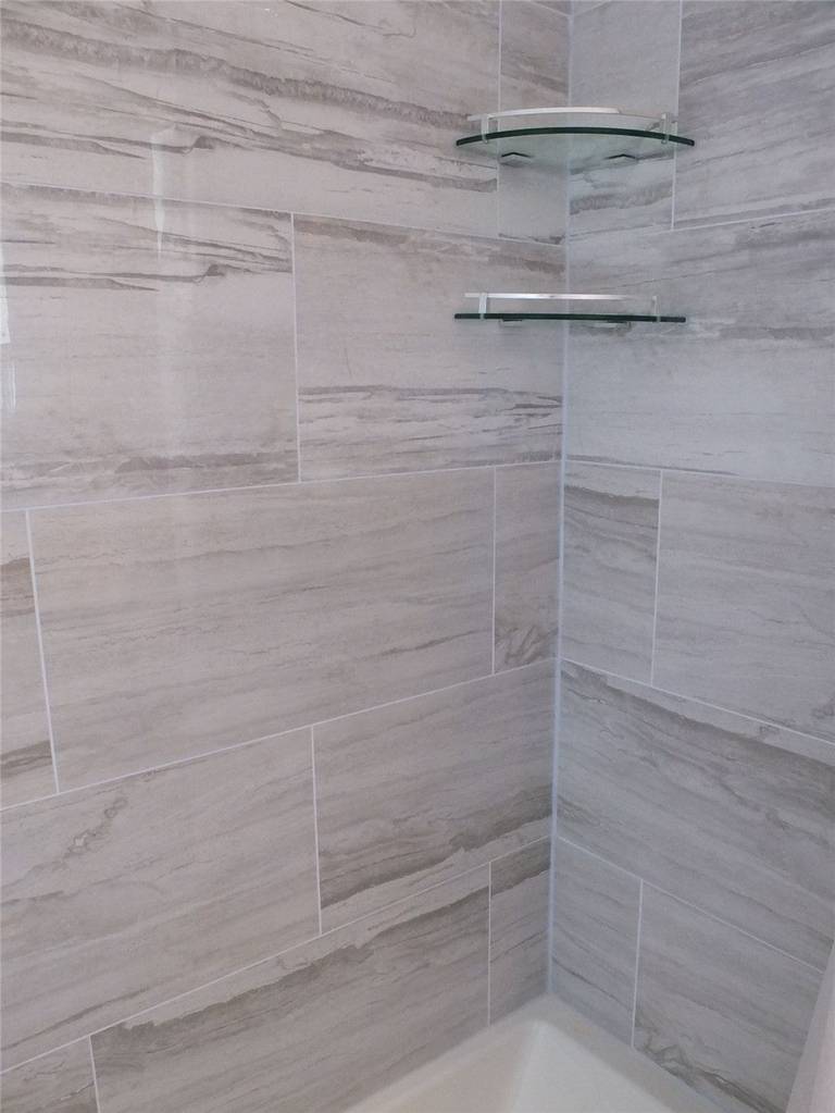This bathroom renovation detail features a shower corner finished with large-format wall tiles and integrated glass shelving for convenient storage. The carefully aligned tile pattern creates visual continuity while maintaining clean grout lines and water-resistant performance. Floating glass shelves add practical organization without interrupting the overall design. Completed by a professional Toronto renovation company, this shower upgrade reflects precise installation and thoughtful space planning.