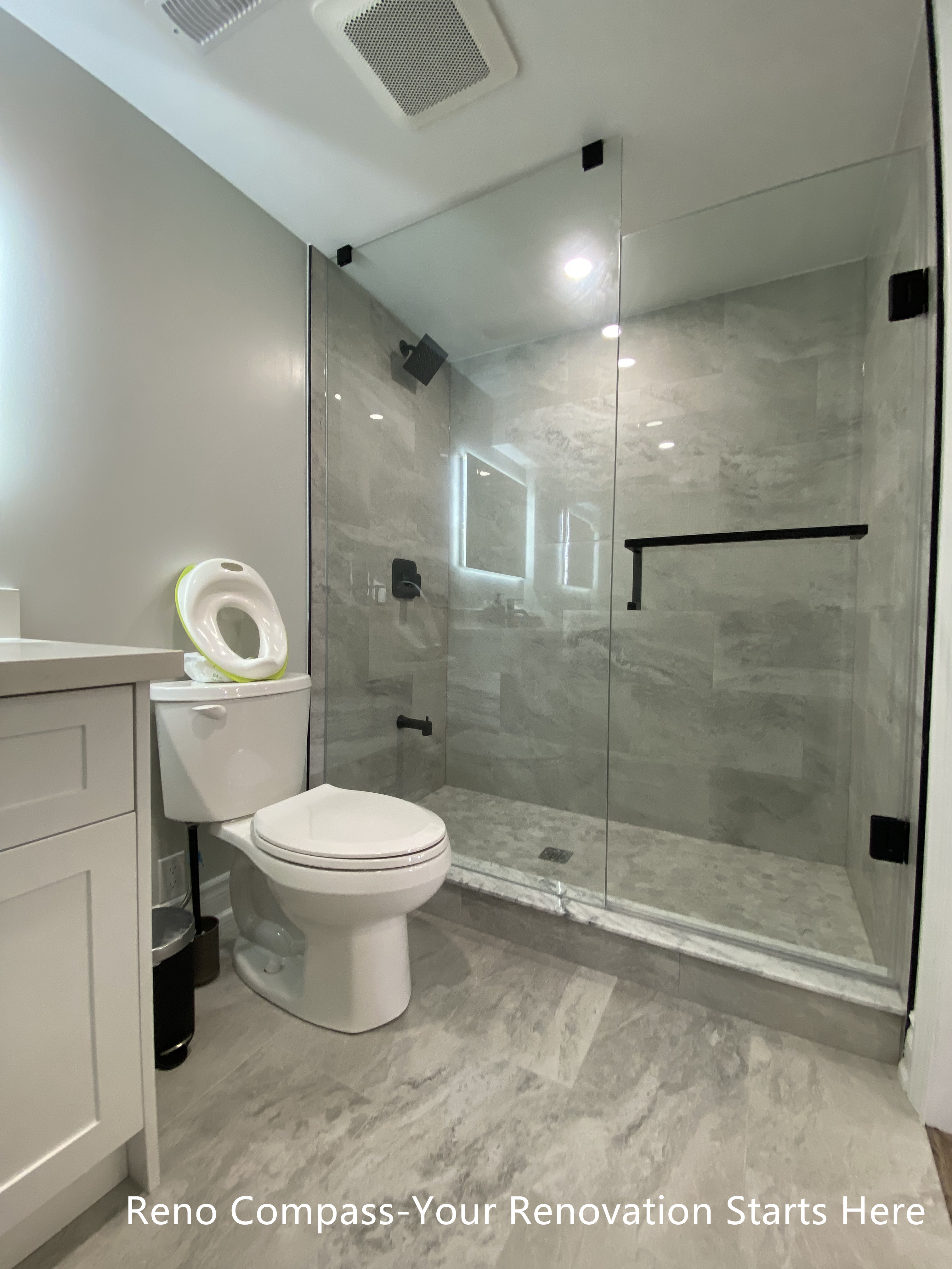 Full view of a modern basement bathroom renovation in Oakville featuring a frameless glass walk-in shower, marble-look tile flooring and walls, matte black shower hardware, efficient ventilation, and a clean, space-saving layout designed for basement living, completed by Reno Compass Inc.