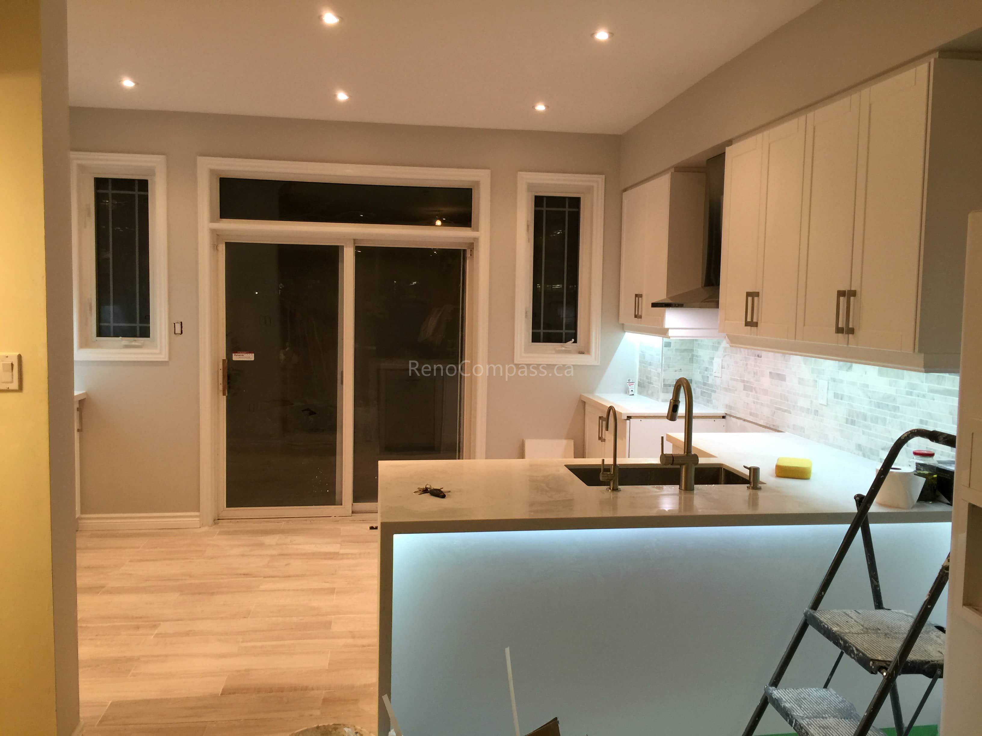 A modern open-concept kitchen interior renovation in Toronto featuring a large central island, integrated LED under-counter lighting, custom cabinetry, and light wood flooring. The design emphasizes functional flow, clean lines, and contemporary finishes, reflecting professional Toronto interior renovation and interior design expertise.