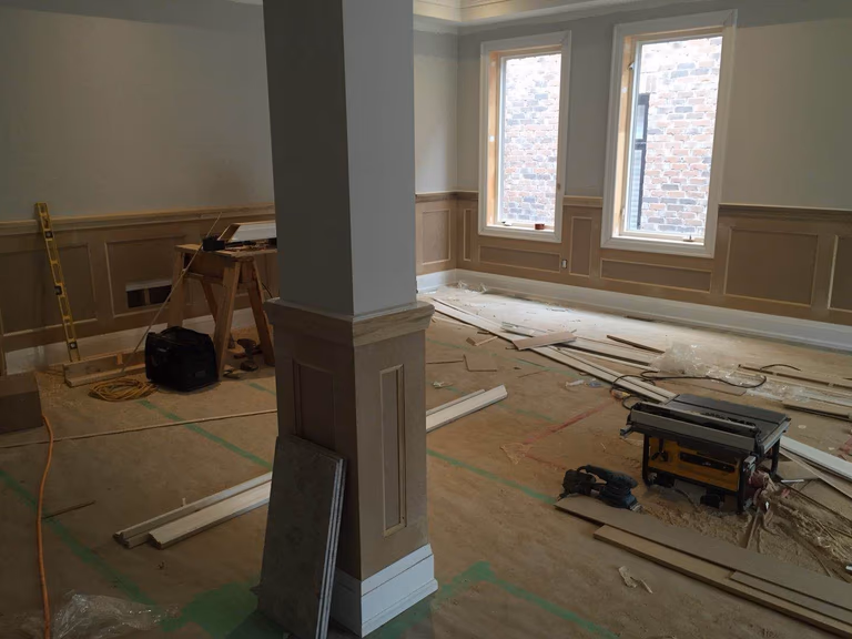 Interior construction phase of a Toronto design build custom build project, showing detailed wall paneling installation and trim work in a bright living area. Craftsmen are preparing decorative wainscoting and interior finishes that add architectural character and depth to the space. This stage is part of a larger whole house renovation Toronto project, where precision carpentry and thoughtful design details contribute to a high-end custom home interior.