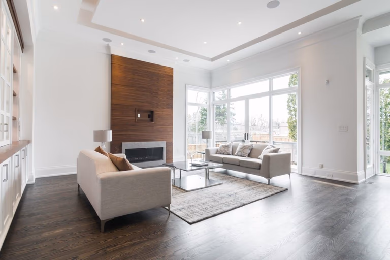 This 4,500 sq ft modern interior renovation in Toronto showcases a bright and expansive living room with floor-to-ceiling windows, a custom wood-paneled fireplace feature wall, and an elegant open-concept layout. The combination of dark hardwood flooring and soft neutral furnishings creates a refined yet inviting atmosphere. Designed and built by a trusted Toronto Renovation Company, Reno Compass Inc., this project reflects premium craftsmanship and sophisticated contemporary living.