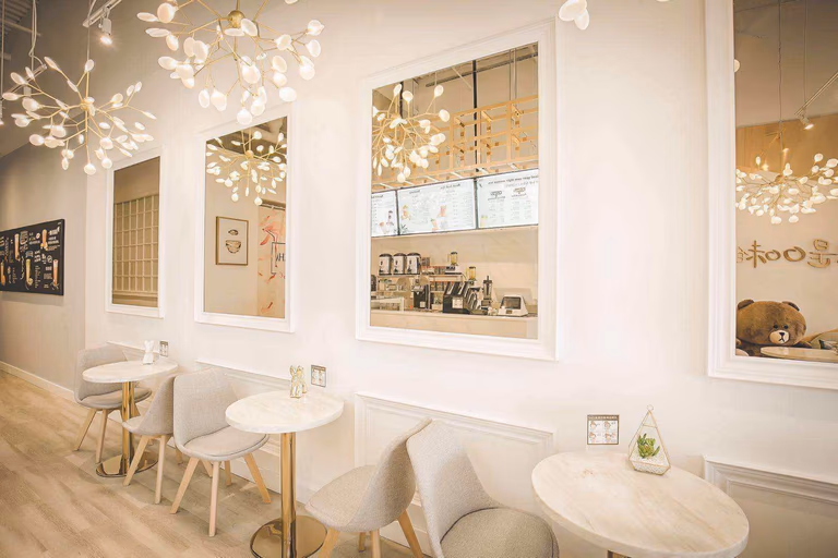 This Toronto bubble tea shop renovation showcases a modern and elegant customer seating area designed to enhance comfort and visual appeal. Featuring custom wall panel detailing, decorative mirrors, warm wood flooring, designer lighting fixtures, and marble-style café tables, the space creates a bright and inviting atmosphere for customers to relax and enjoy their drinks. The layout maximizes seating capacity while maintaining smooth customer flow and an upscale café experience. Completed by a professional Toronto renovation company, this commercial interior project highlights quality craftsmanship, functional space planning, and stylish design tailored for high-traffic milk tea and dessert shop environments.