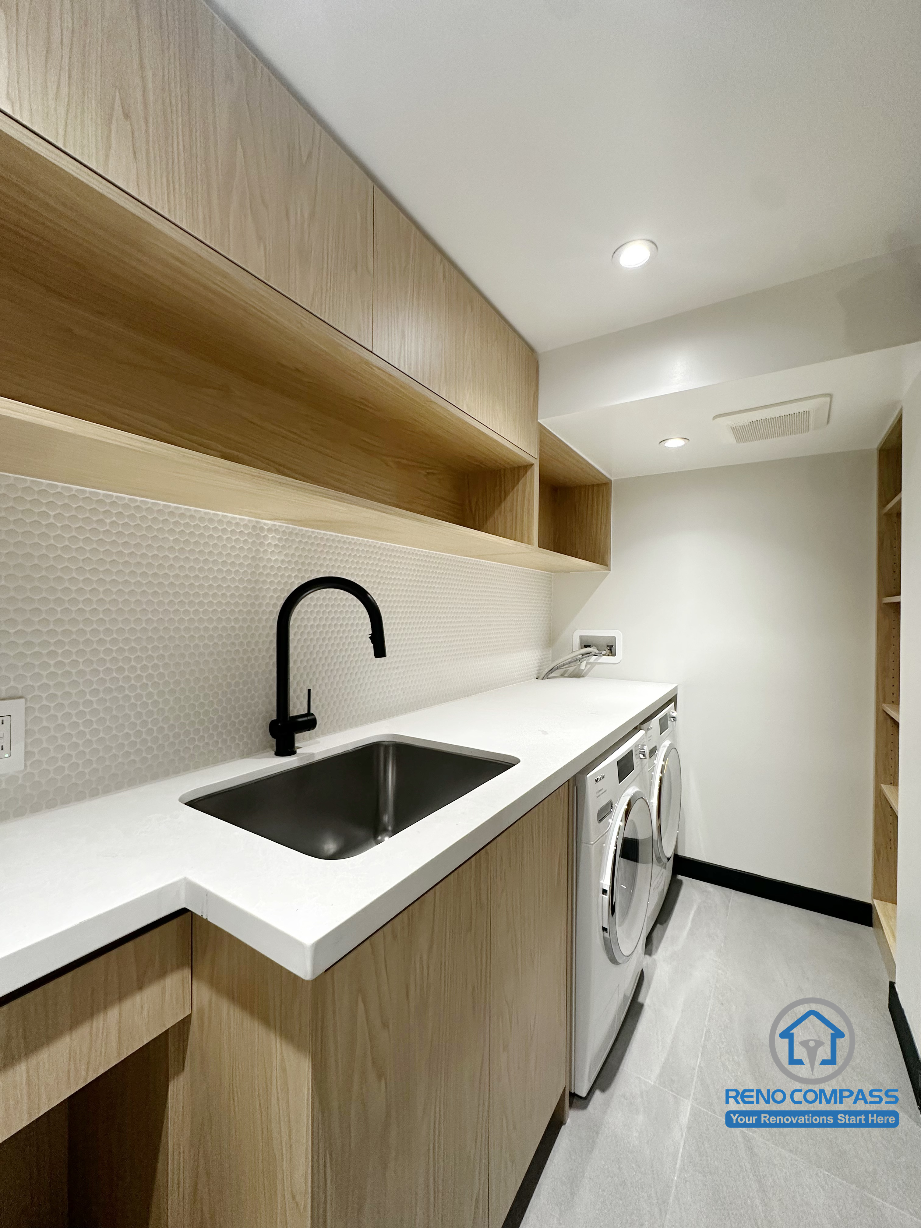 This laundry room renovation in Richmond Hill features custom wood-finish cabinetry, a seamless integrated countertop, and a deep utility sink with matte black faucet. Open shelving and built-in storage enhance organization and workflow, while recessed lighting keeps the space bright and functional. Designed and completed by Reno Compass, this project reflects a clean, modern approach to utility space renovation in the Greater Toronto Area.
