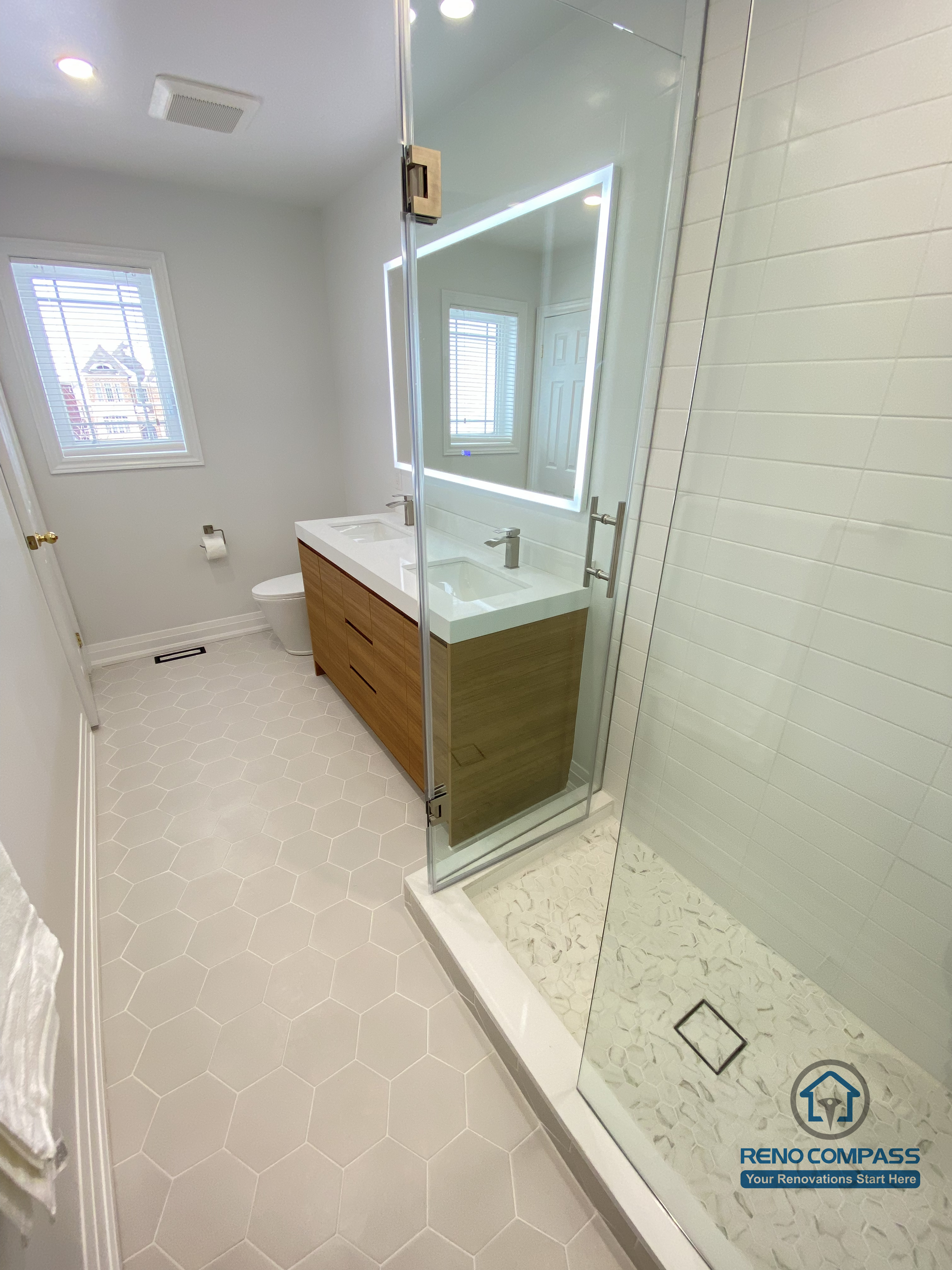 This Markham bathroom renovation showcases an integrated view of the walk-in glass shower and custom vanity, highlighting efficient space planning and clean visual continuity. The frameless glass shower enclosure improves light flow and creates a more open feel, while the tiled shower base and proper drainage ensure safety and durability in daily use. A custom wood-tone vanity paired with a sleek countertop and LED mirror adds warmth and functional storage without compromising simplicity. Neutral finishes and slip-resistant hex tile flooring complete the space with a calm, modern aesthetic. Completed by a Toronto renovation company, Reno Compass Inc., this bathroom renovation reflects a balanced approach to functionality, material coordination, and long-term livability for Markham homes.