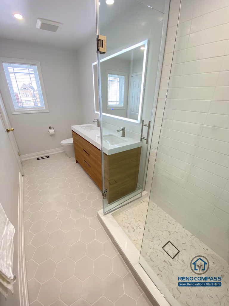 This Markham bathroom renovation showcases an integrated view of the walk-in glass shower and custom vanity, highlighting efficient space planning and clean visual continuity. The frameless glass shower enclosure improves light flow and creates a more open feel, while the tiled shower base and proper drainage ensure safety and durability in daily use. A custom wood-tone vanity paired with a sleek countertop and LED mirror adds warmth and functional storage without compromising simplicity. Neutral finishes and slip-resistant hex tile flooring complete the space with a calm, modern aesthetic. Completed by a Toronto renovation company, Reno Compass Inc., this bathroom renovation reflects a balanced approach to functionality, material coordination, and long-term livability for Markham homes.
