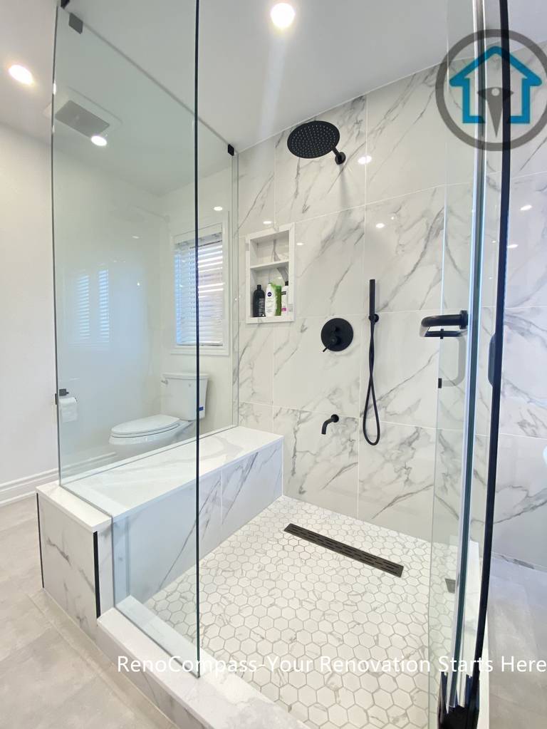 Contemporary Oakville bathroom renovation showcasing a frameless glass walk-in shower, marble-look wall tiles, matte black rain shower system, and hexagon mosaic shower floor. This bathroom remodel highlights precise detailing, clean drainage design, and a modern spa-style layout completed by an experienced Toronto renovation company.