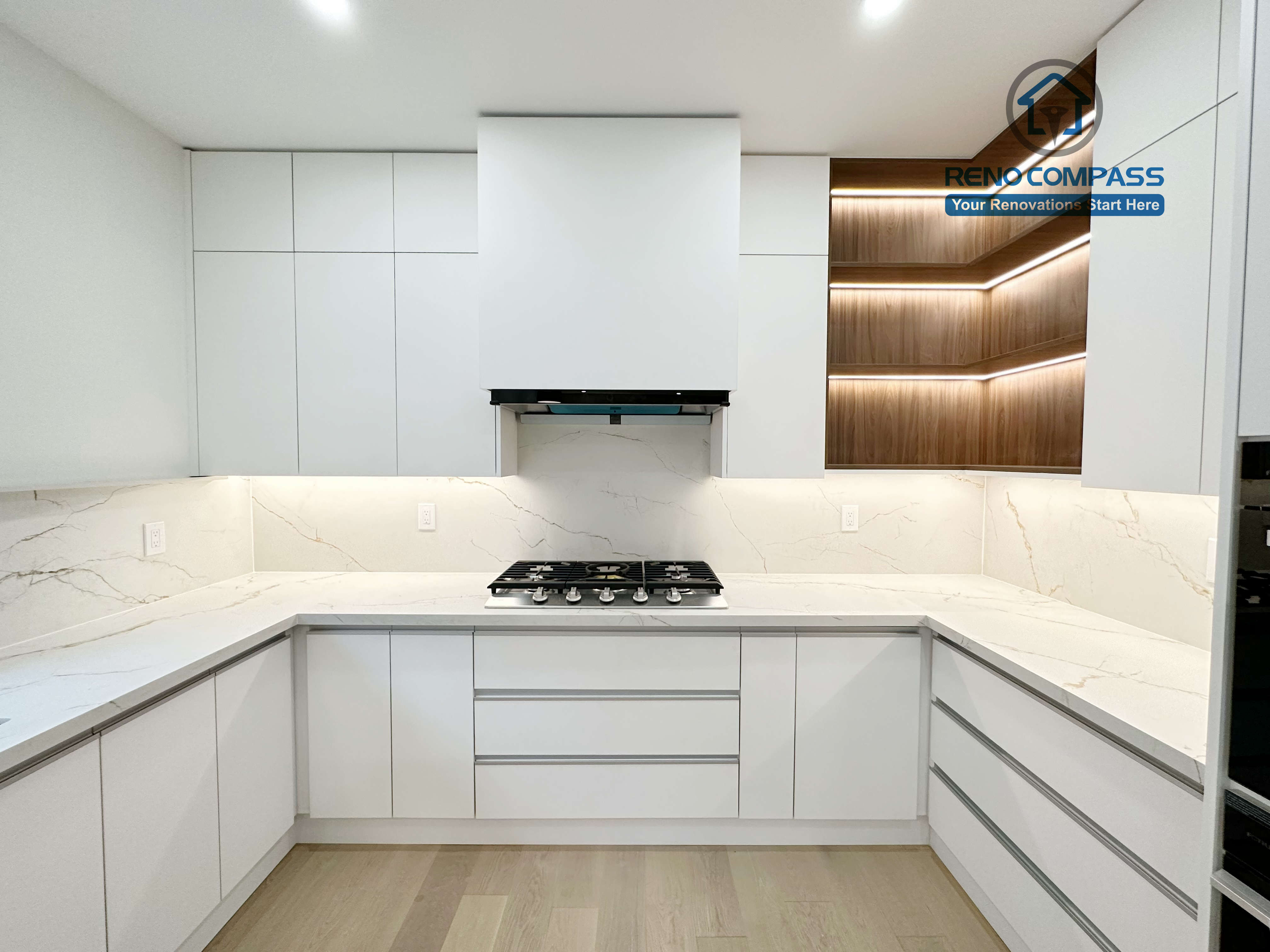 This image showcases a modern U-shaped kitchen renovation in Richmond Hill, featuring full-height custom white cabinetry, a centrally positioned gas cooktop, and seamless quartz countertops with a marble-look backsplash. The clean, handle-less cabinet design maximizes storage and workflow efficiency, while recessed ceiling lighting provides even illumination throughout the space. Warm wood accent shelving with integrated LED lighting adds contrast and depth. Completed by a professional Toronto Renovation Company, this kitchen renovation emphasizes smart layout planning, functionality, and contemporary craftsmanship.
